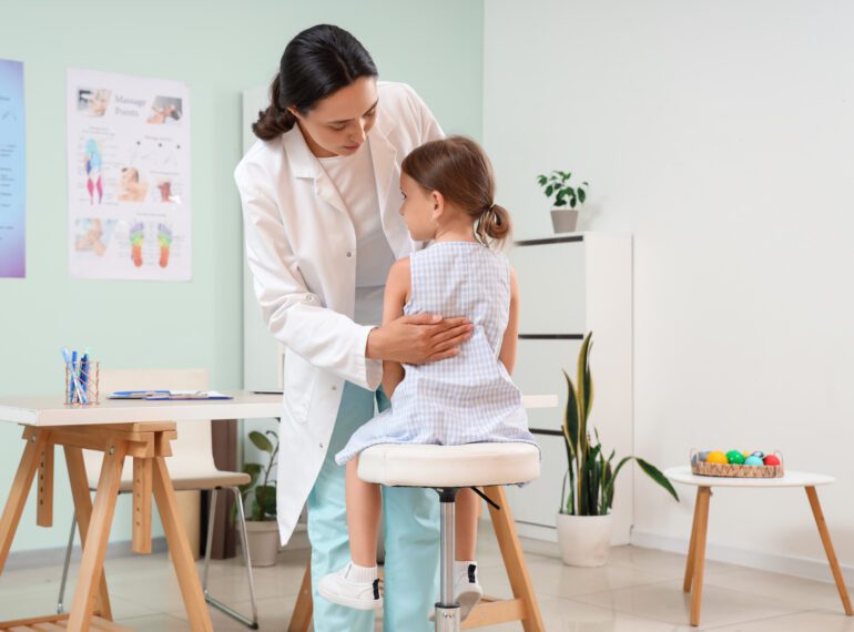 Lekarz w białym fartuchu bada dziecko siedzące na stołku w gabinecie lekarskim.